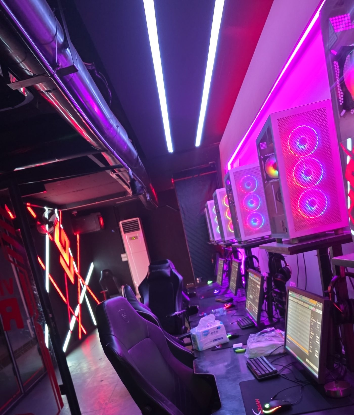 Gaming lounge aisle with white illuminated PC cases on raised shelves and racing seats along the desk