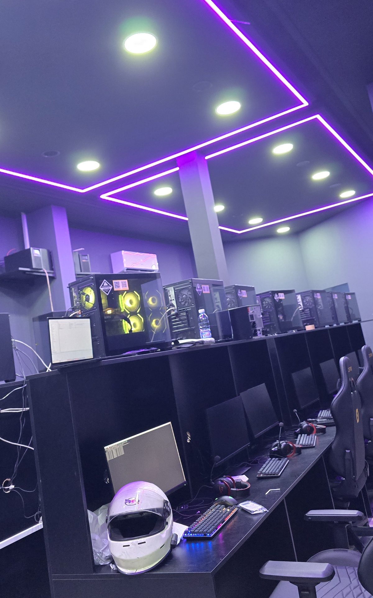 Gaming lounge with wall-mounted tower PCs above a long black desk run, lit by recessed purple ceiling channels