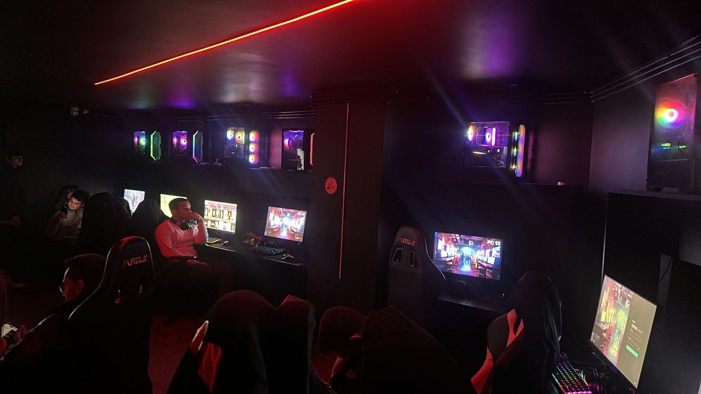 Dark gaming lounge in session, players seated in racing chairs with illuminated PCs mounted on the wall above