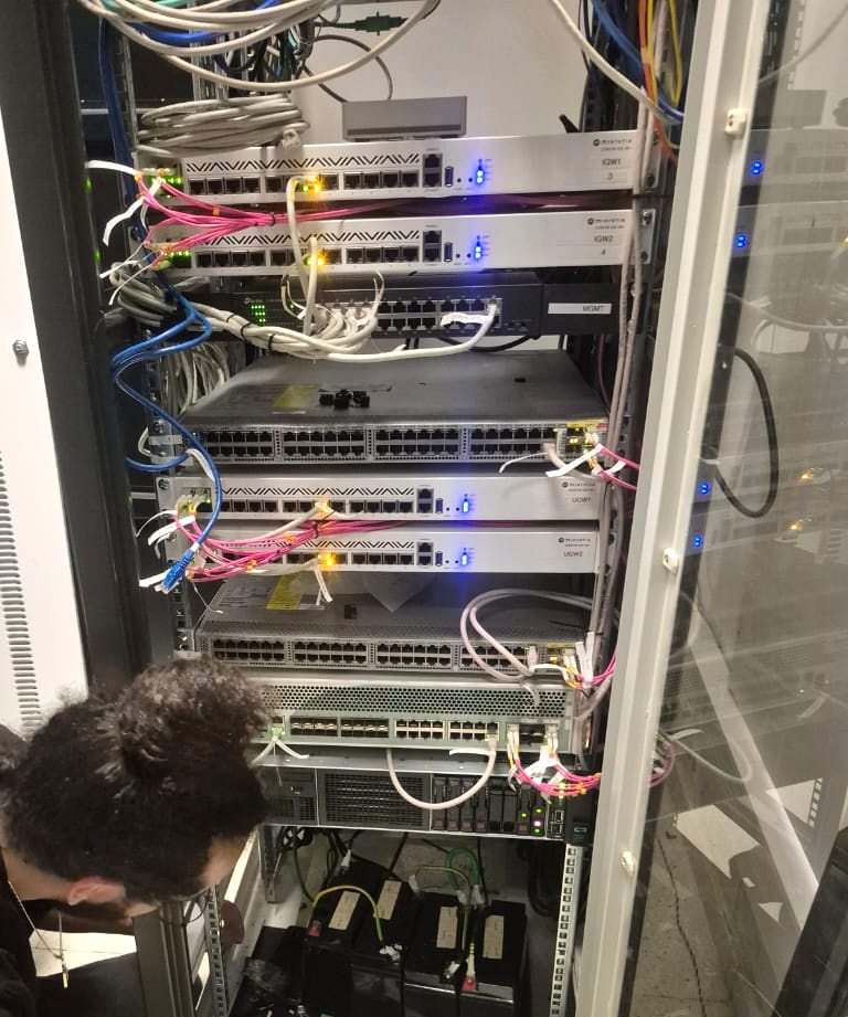 Equipment cabinet with stacked MikroTik routers and switches, labelled gateway units and coloured patch leads, with battery backup below